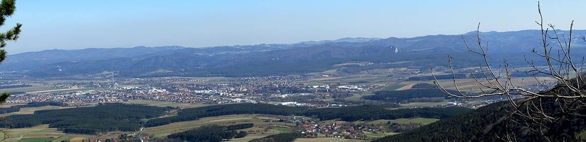 Luftaufnahme einer Stadt umgeben von grünen Hügeln und Bäumen. Gebäude, Felder und eine kurvenreiche Straße sichtbar. Berge in der Ferne unter einem klaren blauen Himmel.