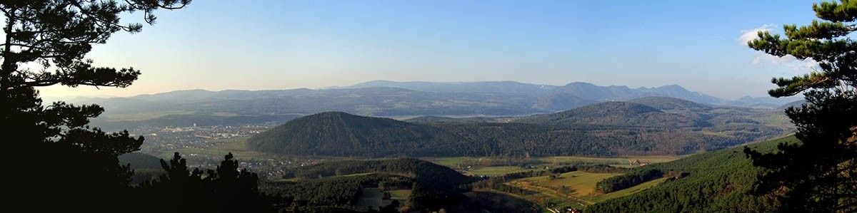 Ein Panoramablick von einem Hügel zeigt ein Tal mit üppigem Grün, eine Stadt in der Ferne und eine Bergkette unter einem klaren blauen Himmel.