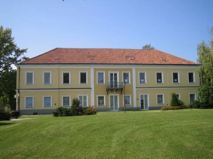 Ein großes gelbes Gebäude mit rotem Dach, vielen Fenstern und einem Balkon steht auf einem großen Rasen, umgeben von Grün und Bäumen unter einem klaren Himmel.