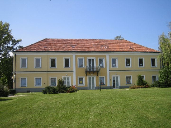 Ein großes gelbes Gebäude mit rotem Dach, vielen Fenstern und einem Balkon steht auf einem großen Rasen, umgeben von Grün und Bäumen unter einem klaren Himmel.