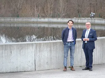 Zwei Männer in Anzügen stehen neben einer Betonmauer an einem Gewässer und lächeln.