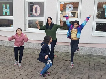 Vier Mädchen und eine Frau, alle lächelnd, springen auf einem Ziegelboden vor einem Gebäude mit großen Fenstern. Die Fenster haben Buchstaben darauf.
