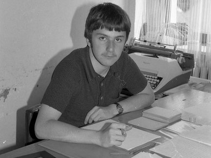 Ein Mann sitzt am Schreibtisch und schreibt, mit einer Schreibmaschine dahinter und mehreren Papieren auf dem Schreibtisch.