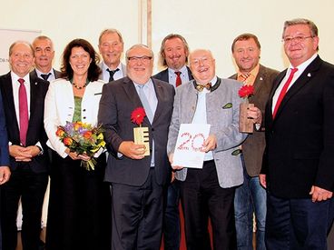 Neun Personen posieren für ein Foto. Sie alle haben ein Lächeln im Gesicht. Jeder von ihnen hält entweder eine Blume oder ein Zertifikat. Sie sind alle formell gekleidet.