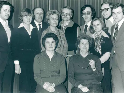 Ein Schwarz-Weiß-Foto einer Gruppe von Menschen, einige stehend und einige sitzend, in formeller Kleidung, lächeln für die Kamera.