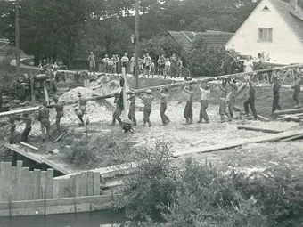 Ein Schwarz-Weiß-Bild einer Gruppe von Menschen, die eine große Holzbalken heben, wahrscheinlich an einer Baustelle. Sie tragen Hüte und Schuhe. Im Hintergrund befinden sich Häuser und Bäume.