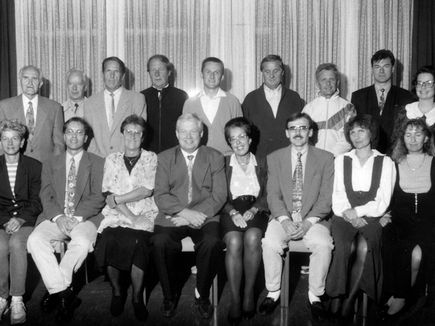 Eine Gruppe von Menschen, sowohl Männer als auch Frauen, gekleidet in formeller Kleidung, posiert für ein Foto. Sie sitzen auf Stühlen und hinter ihnen befinden sich weiße Vorhänge.