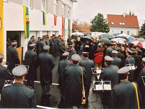 Eine Gruppe von Menschen in Uniform, möglicherweise Militärangehörige, steht vor einem Gebäude mit Fahnen. Sie halten Musikinstrumente und Regenschirme, möglicherweise bei einer Aufführung.