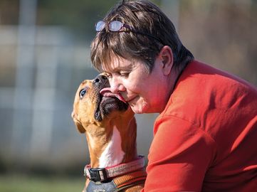 Eine Frau in einem roten Shirt ist draußen mit einem braun-weißen Hund, der ein Halsband trägt. Sie küsst die Nase des Hundes.