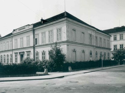 Ein Schwarz-Weiß-Bild zeigt ein großes Gebäude mit mehreren Fenstern und einem zentralen Eingang, beschriftet mit 'Volksschule.' Das Gebäude wird von Hecken und Bäumen flankiert.