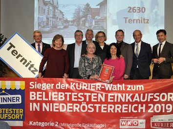 Eine Gruppe von Menschen steht hinter einem Banner und hält ein rotes Buch. Sie sind in formeller Kleidung und scheinen zu feiern. Der Schriftzug auf dem Banner lautet 'SIEGER DER KURIER-WAHL ZUM BELIEBTESTEN EINKAUFSORT IN NIEDEROESTERREICH 2019'.