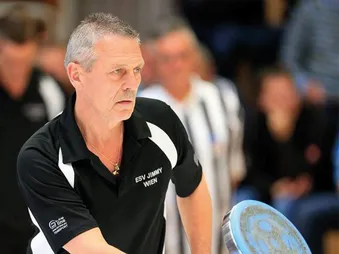 Ein Mann in einem schwarzen Shirt spielt Darts, mit einem unscharfen Hintergrund von einer Menschenmenge.