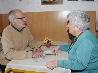 Ein älteres Paar sitzt an einem Tisch und führt ein Gespräch. Der Mann liest von einem Papier. Der Tisch hat eine kleine Topfpflanze.