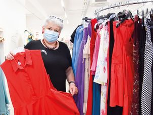 Eine Frau mit Maske hält ein rotes Oberteil in einem Bekleidungsgeschäft mit verschiedenen Kleidern an Kleiderbügeln.