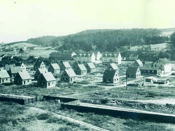Ein Luftbild einer Stadt mit mehreren Häusern und Gebäuden. Die Stadt ist von Bäumen umgeben und hat eine Eisenbahn im Vordergrund.