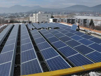 Ein Dach, das mit Solarmodulen bedeckt ist, und ein Mann, der ein Panel hält. Im Hintergrund befinden sich Gebäude und Berge.