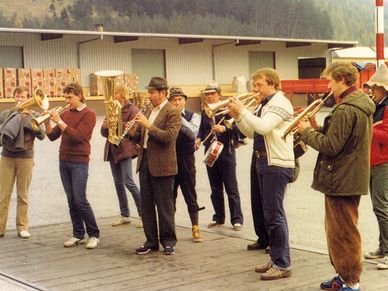 Eine Gruppe von Musikern spielt Trompeten, Tubas und Trommeln auf einer Holzplattform vor einem Gebäude.