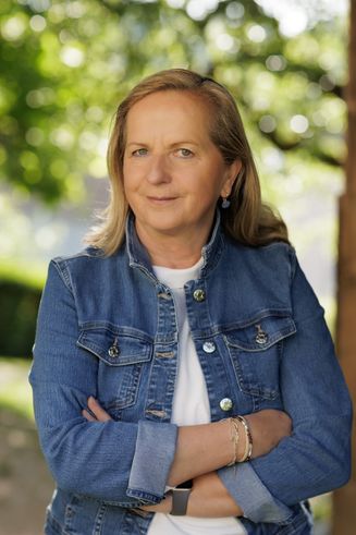 Eine Frau mit blonden Haaren, die eine Jeansjacke und ein weißes T-Shirt trägt, lächelt mit verschränkten Armen.