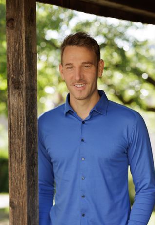 Ein Mann in einem blauen Hemd lächelt und steht draußen in der Nähe eines Holzpfostens.