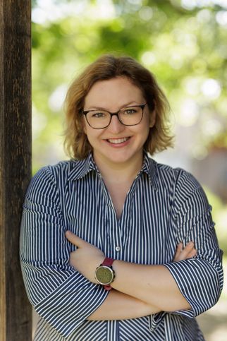 Eine Frau mit kurzen blonden Haaren, die eine Brille und ein gestreiftes Hemd trägt, lächelt und posiert für ein Foto.