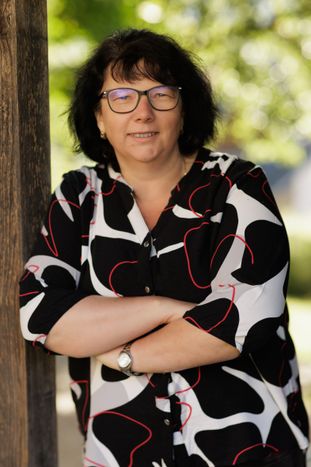 Eine Frau mit kurzen schwarzen Haaren, die eine Brille trägt und ein gemustertes schwarz-weißes Hemd trägt, steht mit verschränkten Armen.
