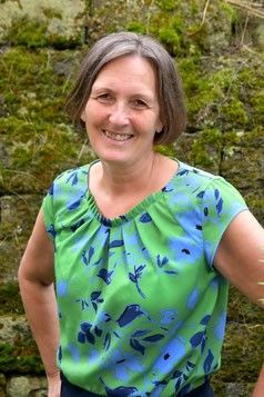 Eine Frau mit kurzen Haaren steht vor einer moosbedeckten Wand und lächelt. Sie trägt ein grün-blaues Oberteil mit Blumenmuster.