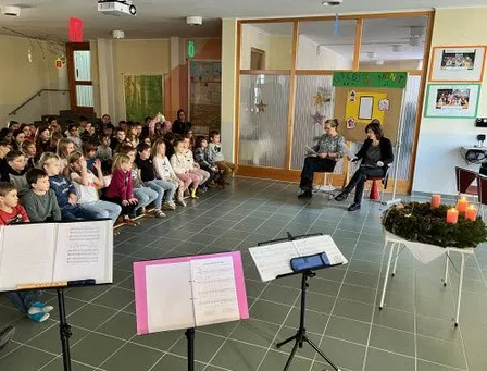 Eine Gruppe von Kindern sitzt auf Bänken mit Blick nach vorne, wo zwei Personen sitzen. Notenblätter sind vor den Kindern auf Ständern platziert. Kerzen stehen auf einem Tisch und eine Weihnachtsdekoration ist auf dem Tisch platziert.