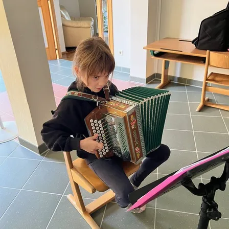 Ein kleines Mädchen sitzt auf einem Stuhl und spielt Akkordeon mit einem Notenständer davor. Hinter ihr ist ein Raum mit einem Holzschreibtisch, einem Stuhl und einer Couch.