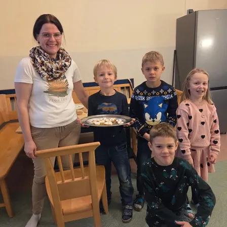 Eine lächelnde Frau mit Brille hält einen Tablett mit Keksen, während vier Kinder neben ihr stehen und für ein Foto in einem Raum posieren.