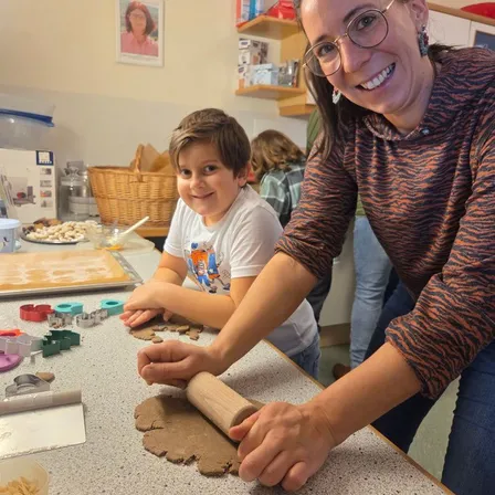 Eine Frau und ein Junge sind in der Küche und backen Kekse mit einem Nudelholz. Der Junge sitzt an einem Tisch, und die Frau steht neben ihm. Es gibt Keksausstecher, ein Tablett mit Keksen und eine Schüssel mit Kekskrümel auf dem Tisch.