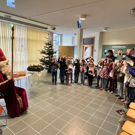 Eine Gruppe von Kindern steht um einen Weihnachtsbaum herum mit einem Mann, der als Weihnachtsmann verkleidet ist. Sie heben ihre Hände. Ein Klavier und ein Stuhl sind auf der linken Seite.