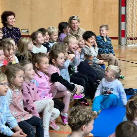 Eine Gruppe von Kindern sitzt im Kreis auf dem Sportboden und beobachtet eine Frau und lacht.