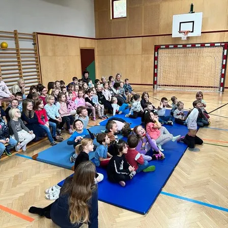 Eine Gruppe von Kindern und Erwachsenen sitzt auf blauen Matten in einer Turnhalle. Einige lachen, während andere etwas betrachten. In der Ferne ist ein Basketballkorb zu sehen.