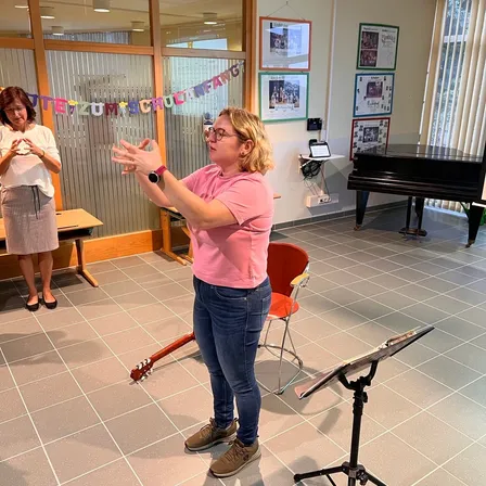 Eine Frau in einem rosa Shirt und Jeans steht in einem Musikraum und gestikuliert mit ihren Händen. Eine Frau in einem weißen Shirt und Rock steht neben ihr. Ein roter Stuhl und ein Notenständer liegen auf dem Boden. Ein Klavier steht in der Ecke des Raumes.