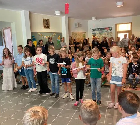 Eine Gruppe von Kindern steht im Kreis, Papiere in der Hand, vielleicht im Proben für ein Schulstück, während Erwachsene und Kinder ihnen in einem Raum zuschauen.