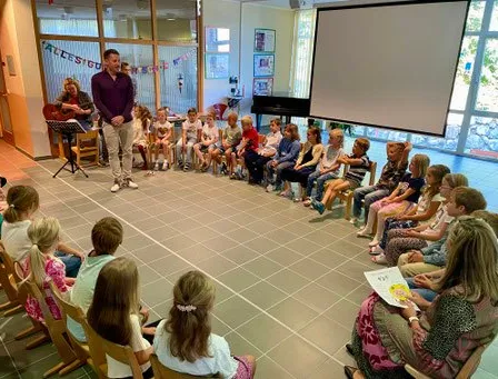 Ein Mann steht vor einem Klassenzimmer voller Kinder und hält eine Gitarre. Die Kinder sitzen auf Stühlen und lauschen. Der Raum hat eine Projektionsleinwand und ein Klavier.