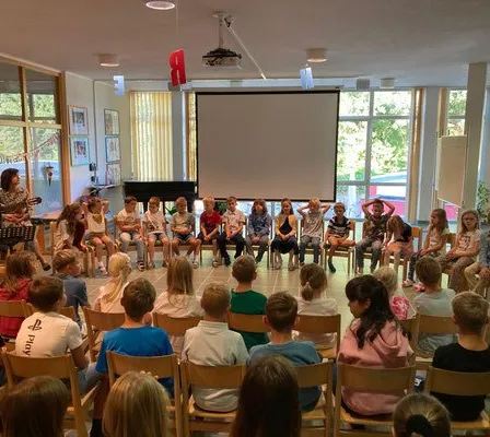 Eine Gruppe von Kindern in einem Klassenzimmer, auf Stühlen sitzend, beobachtet einen Erwachsenen, der eine Gitarre spielt. Ein Beamerbildschirm ist im Hintergrund, und es gibt Glasfenster mit Vorhängen.