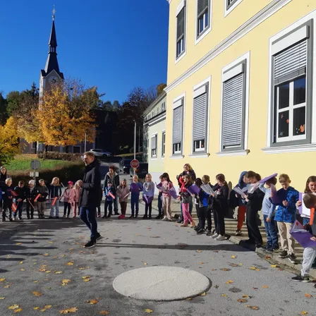 Ein Mann führt eine Reihe von Kindern vor einem Gebäude an, hält Papiere in der Hand, auf einer Straße mit Herbstblättern und einer Kirche im Hintergrund.