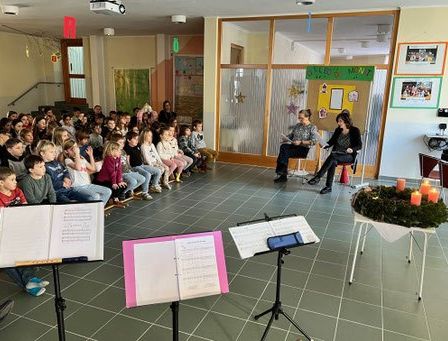 Eine Gruppe von Kindern sitzt auf Bänken mit Blick nach vorne, wo zwei Personen sitzen. Notenblätter sind vor den Kindern auf Ständern platziert. Kerzen stehen auf einem Tisch und eine Weihnachtsdekoration ist auf dem Tisch platziert.