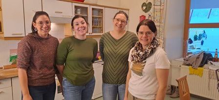 Fünf Frauen stehen in einer Küche und lächeln. Sie tragen verschiedenfarbige Oberteile und Brillen. Eine trägt einen Schal. Hinter ihnen ein Fenster mit Blick nach draußen.
