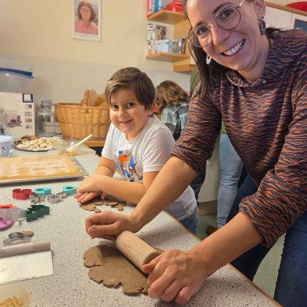 Eine Frau und ein Junge sind in der Küche und backen Kekse mit einem Nudelholz. Der Junge sitzt an einem Tisch, und die Frau steht neben ihm. Es gibt Keksausstecher, ein Tablett mit Keksen und eine Schüssel mit Kekskrümel auf dem Tisch.