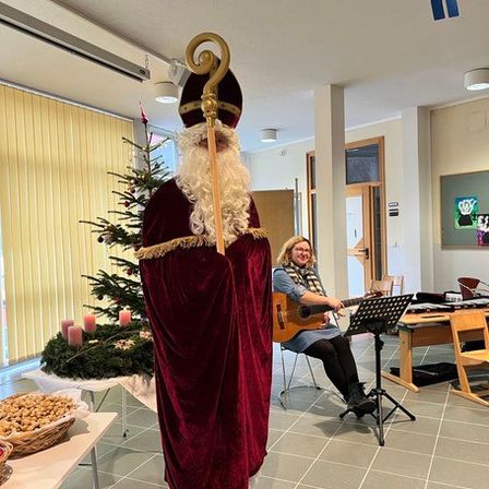 Eine Person in einer Weihnachtsmann-Verkleidung steht mit einem Gehstock in einem Raum mit einem Weihnachtsbaum und einer Frau, die Gitarre spielt.