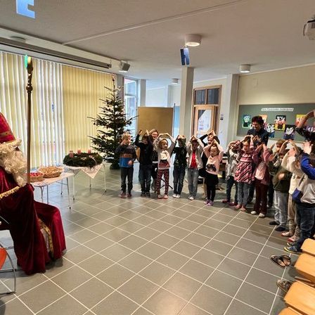 Eine Gruppe von Kindern steht um einen Weihnachtsbaum herum mit einem Mann, der als Weihnachtsmann verkleidet ist. Sie heben ihre Hände. Ein Klavier und ein Stuhl sind auf der linken Seite.