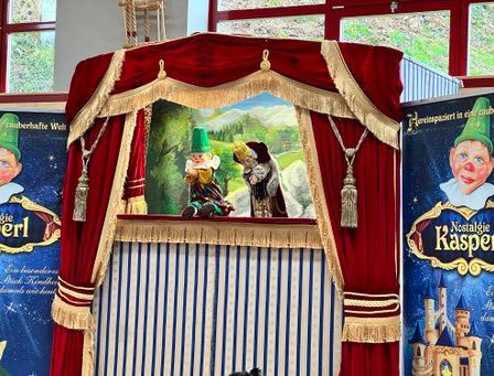 Eine Puppentheater-Ausstellung mit rotem und goldenem Vorhang im Hintergrund, bei der zwei Zwerge mit einer Landschaftsmalerei hinter ihnen zu sehen sind. An den Seiten sind Werbungen für eine Show.