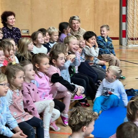 Eine Gruppe von Kindern sitzt im Kreis auf dem Sportboden und beobachtet eine Frau und lacht.