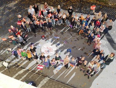 Eine Gruppe von Kindern und Erwachsenen steht im Freien im Kreis und hält Fahnen und Plakate. Der Boden ist mit Blättern bedeckt.