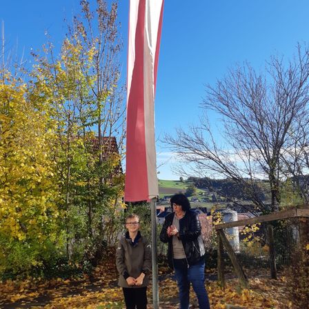 Zwei Personen stehen vor einem Fahnenmast in einer Außenumgebung mit Bäumen und einem Haus im Hintergrund.