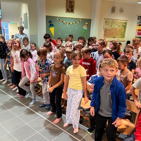 Kinder in einem Klassenzimmer mit einer Frau vor einer Tafel und einer Karte. Sie tragen Hausschuhe und manche lächeln.