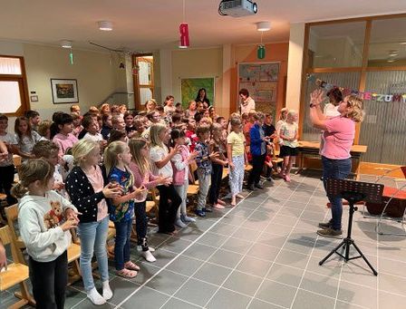 Eine Gruppe von Kindern ist in einem Raum versammelt, trägt Freizeitkleidung und steht in einer Reihe. Eine Frau steht vor ihnen mit einem Mikrofon auf einem Ständer. Sie hält ein Kind auf der Schulter und scheint zu singen. Der Raum hat Holzstühle, Fliesenboden und einen an der Decke montierten Projektor.
