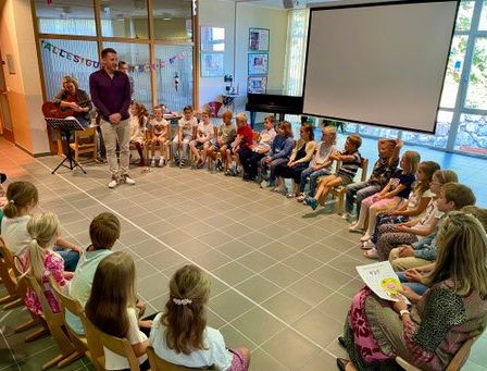 Ein Mann steht vor einem Klassenzimmer voller Kinder und hält eine Gitarre. Die Kinder sitzen auf Stühlen und lauschen. Der Raum hat eine Projektionsleinwand und ein Klavier.